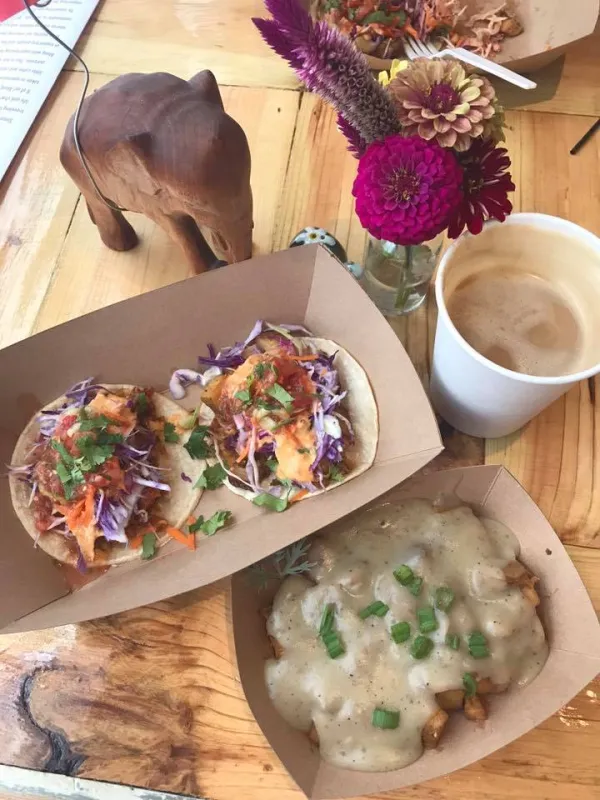 A top down view of a meal - a brown holder with what looks like two taco-like things, and brown drink in a white cup, something in a brown holder at the bottom, white potatoes with something green sprinkled on top and a red flower in a vase and an elephant statue