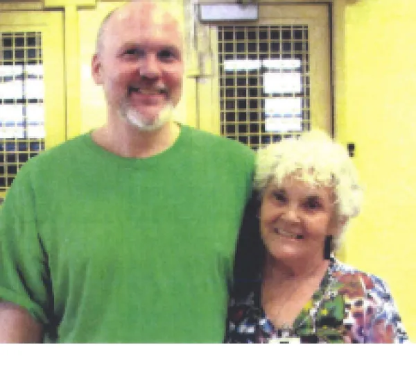 Tall bald white man with a mustache and beard and green T-shirt standing smiling with his arm around a gray haired smiling lady in a flowered dress