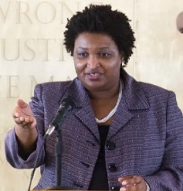 Black woman in a gray suit with a white necklace making a gesture while talking at a microphone