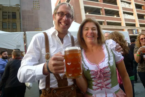 White ma with gray hair and glasses smiling holding a huge mug of beer with a woman who is wearing a German-looking corset and frilly top