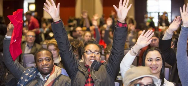 Black woman with glasses in front of a huge crowd of people smiling with both hands held high above her head
