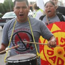 Latino man in foreground with drum around his neck and hitting it with drumsticks, his mouth open chanting, wearing a shirt that says Boycott Wendys with a picture of a girl's head with red pigtails and a No sign around it (circle with line through it) and a Latino woman behind him holding a bright orange and yellow sign also with her mouth open chanting