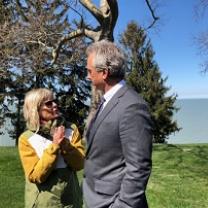 Short blonde wtoman wearing a yellow coat and sunglasses talking and gesturing to a taller white man with gray hair and a gray suit outside by tree and a lake 