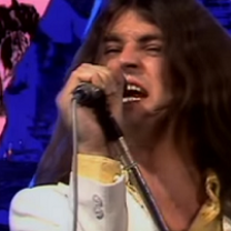 Long white man rock singer belting out a song into a mic with a blue and pink psychedelic background