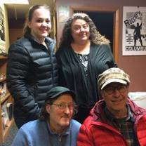 Two women, one young with hair pulled back and the other older with long brown curly hair standing behind two men kneeling, both wearing hats with brims and wire rimmed glasses, everyone smiling