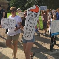 People marching outside in shorts with trees in background carrying signs, one saying Give us your tired poor