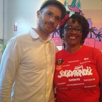 Young white man with facial hair, glasses and a hat posing with his arm around a black woman in a solidarity T-shirt