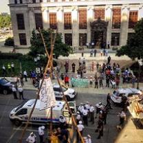 Scene in downtown with tall office buildings lots of people in the streets with protest signs, a lot of police and a tepee looking wooden structure