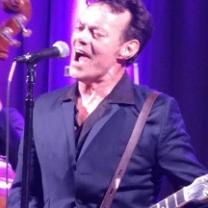 White man at a microphone belting out a song wearing a dark suit and shirt unbuttoned with a guitar strapped around him