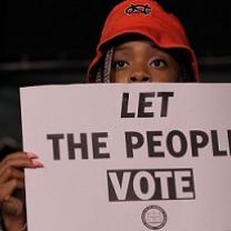 Black person's face with a red hat on their head peering over a sign that they are holding that says Let The People Vote