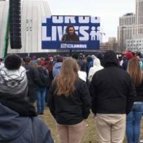 Backs of people standing watching a speaker on a screen