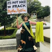 Woman in a long green dress with a sash and a cape and crown of flowers with a sign saying It's not nice to frack mother nature