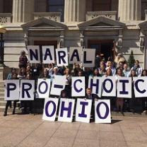 Big white capitol building with columns and turrets above windows in the background, lots of people posing in front holding placards each with a letter spelling out NARAL PRO-CHOICE OHIO