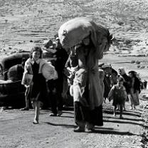 Black and white photo of women and children walking down a dusty path outside with the woman in the foreground carrying a big heavy bag on her head