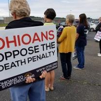The backs of four people at a vigil outside one holding a sign that says Ohioans Oppose the Death Penalty
