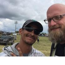 Bearded white man with glasses an bald head smiling with a dark-skinned man in sunglasses and a baseball cap outside in front of some cars and a cloudy sky