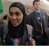 Young dark skinned woman in a burqa smiling and a young white man in a suit looking serious behind her