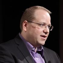 Middle aged white man with brown receding hair with wire rimmed glasses looking to the right with  a gray suitcoat and purple shirt his mouth open as if talking