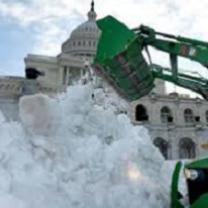 Big white stately building with round top and lots of windows behind a huge green truck with a big arm and huge shovel putting a ton of white snow in a big pile in front
