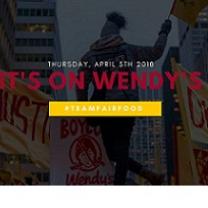 Darkened photo of back of a black woman outside waving a yellow flag over her head and signs all around her about justice and against Wendys