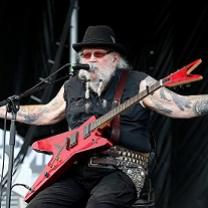 Older man with black hat, white hair and beard, sunglasses, sitting on a chair on stage with his arms spread wide with lots of tattoos and a red rock and roll electric guitar on his lap