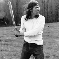 Black and white photo of a young man with long flowing brown hair with a headband across his forehead holding a baseball bat as if he's about to swing