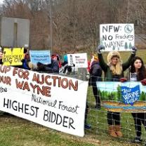 Lots of young people wearing coats outside near a forest at a fence with protest signs reading Up for Auction Our Wayne to the Highest Bidder