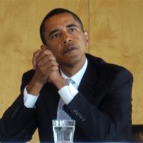 Obama with elbows on table clasping his hands together looking pensive