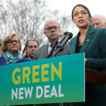 Sign saying Green New Deal with people behind it and young woman with glasses speaking
