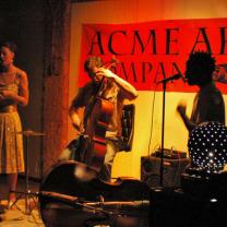 A red sign sayigng ACME ART on the wall behind silhouettes of musicians, a guy playing a bass fiddle, a person singing into a mic and a woman wearing a mid-length dress