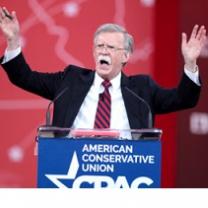 Older man with gray hair and long white mustache standing at a podium with his mouth  open waving both hands in the air and the words American Conservative Union on the podium