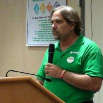 White man with goatee and dark hair in green shirt holding a mic at the podium