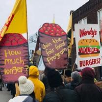 People marching with very large high signs with depictions of barrels of tomatoes with the words How much longer? and Join the Fair Food program and a Wendy's hamburger that says violence on it