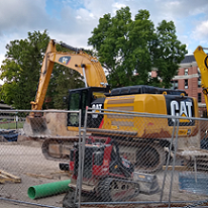 Big yellow machine truck with big scoop on a long neck that says CAT on the side behind a wire fence with other machinery and trees in the background