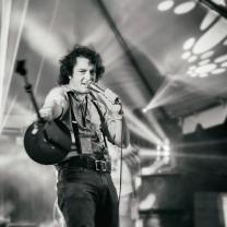 Black and white photo of young dark haired man singing into a mic on a stage