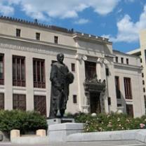 Big governmental building with statue of Christopher Columbus in front, some bushes against a blue sky with white clouds
