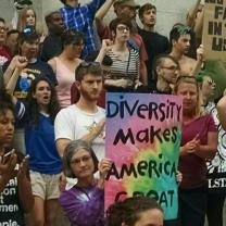 Person holding sign in crowd of people with fists in air, reading Diversity Makes America Great against tie-dyed background