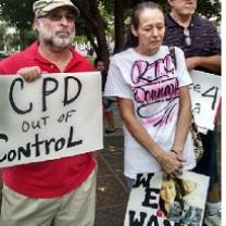 Man with gray beard and sunglasses wearing a baseball cap holding a sign that says CPD out of control standing next to an older woman with gray hair pulled back in a ponytail looking down at the ground, very sad, wearing a white T-shirt with words "RIP Donna" on it and holding a sign with pictures of a young woman on it