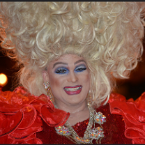 Man dressed a woman with huge blonde hair in curls going up in the air lots of blue eye shadow, red lips and a red outfit