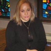Young Latina woman with blonde hair and black clothes sitting on a pew in a church with stained glass windows behind her