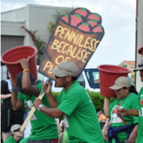 Dark-skinned men and women in green shirts at a demonstration marching to the left one holding a sign saying Penniless because of and two others holding big rust colored buckets