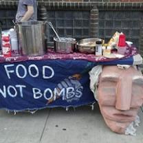 Big paper mache face hanging on the front of a table outside next to a blue sign with white hand drawn letters that say Food Not Bombs and a lot of pots and pans and condiments on the table and a brick wall in the background