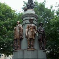 Gray pointy statue with Union Soldier figures standing with their backs around it against trees in the background