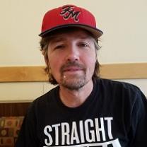 Man with brown hair sticking out from a red baseball cap and goatee wearing a black T-shirt 