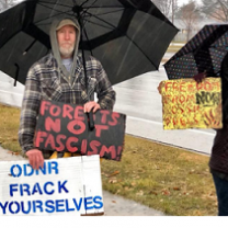 Two people standing outside in the rain holding umbrellas and signs, one says ODNR Frack Yourselves