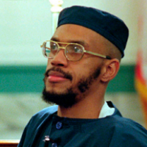 Black man with wire rimmed glasses and mustache and beard and a square topped blue hat