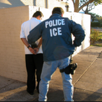 Back of man in blue jacket with white letters reading ICE putting another man with his back to us in handcuffs