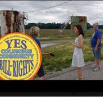 People standing outside in front of weird looking rusty metal structures and the round yellow logo for Columbus Community Bill of Rights