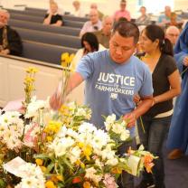 Main in a Justice for Farmworkers light blue T-shirt putting flowers into a huge container that has a lot of flowers in it and a line of people behind him and pews of a church behind them