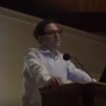 White man with glasses and white shirt standing and speaking at podium
